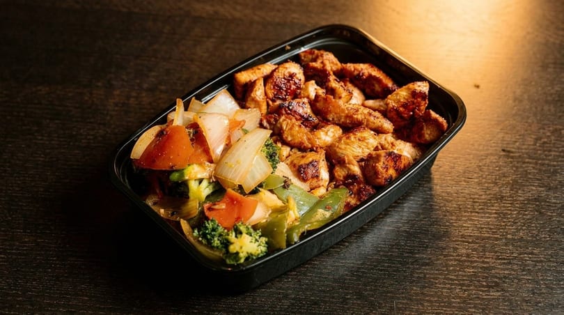 Grilled chicken and stir-fried vegetables in a black takeout container on a dark wooden table.