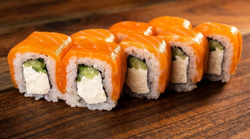 Eight salmon sushi rolls with cream cheese and cucumber on a wooden surface.