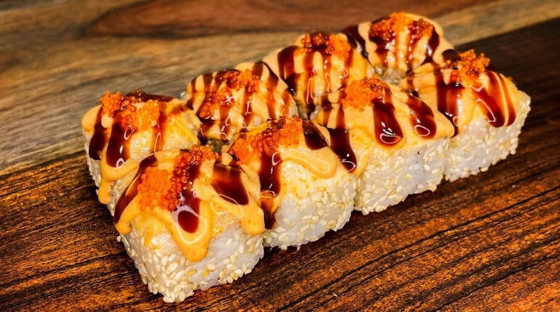 Eight sushi rolls with sesame seeds, drizzled with orange sauce, dark glaze, and orange tobiko.