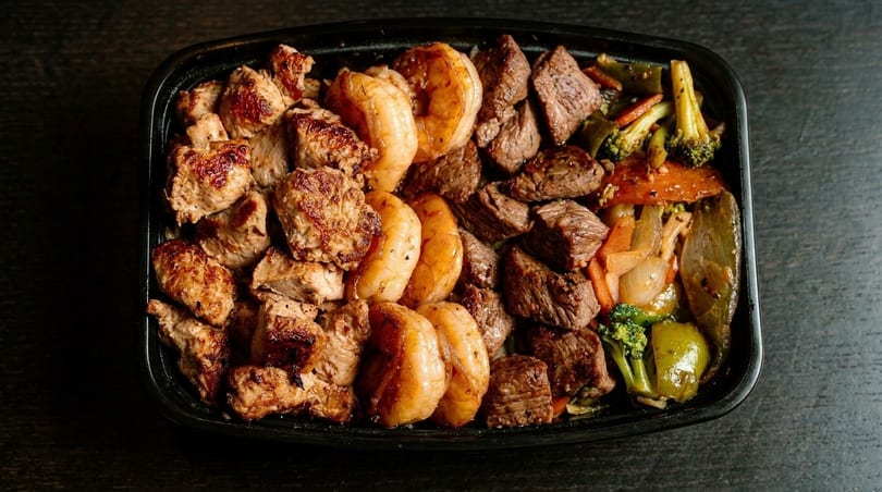 A black container filled with grilled chicken, shrimp, steak, and mixed vegetables.
