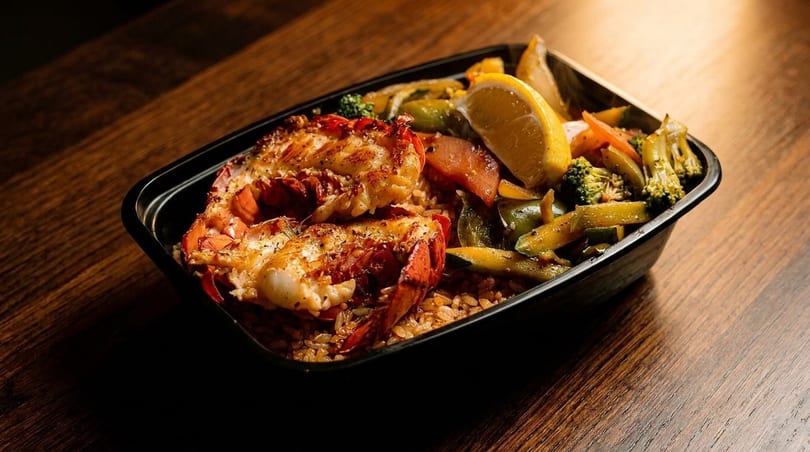 Grilled lobster tails with rice, mixed vegetables, and a lemon wedge in a black container.