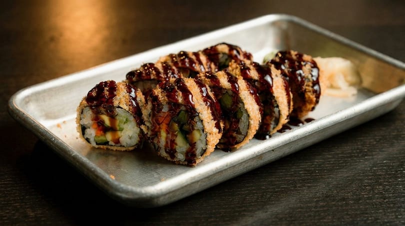 Crispy fried sushi rolls drizzled with dark sauce, served on a metal tray with pickled ginger.